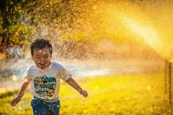 A little boy running with a smile on his face while a sprinkler is spraying water in his direction. The drops of water are golden because the sun is hitting them