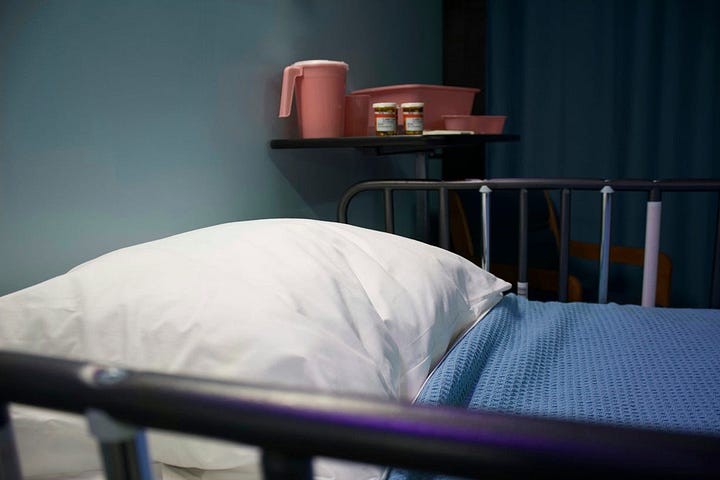 An attractive view of a made-up hospital or nursing home bed, similar to the one my sister had in her room. The shelf in the background has the added touch of having the water pitcher and the prescription bottles sitting there.
