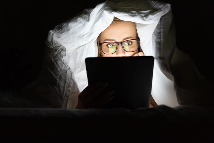 Woman in glasses lying in bed under the blanket using tablet— doomscrolling late at night.