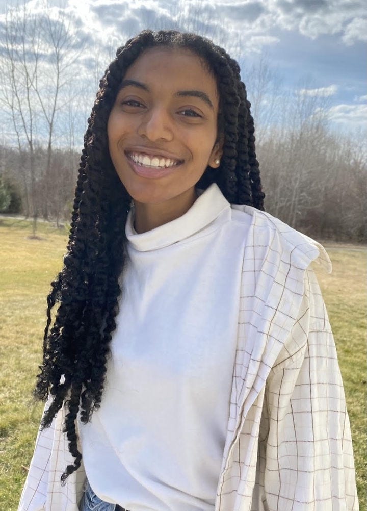 Image of young Black woman with long, dark hair in twists, smiling and wearing a white turtleneck sweater and off-white long-sleeved shirt.