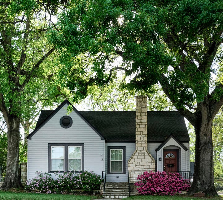 An older home set between large trees. It’s while with grey trim and a dark wooden door, with a light beige brick chimney.