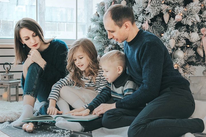 A small girl and boy reading a book, sitting under a Christmas tree, between a man and woman