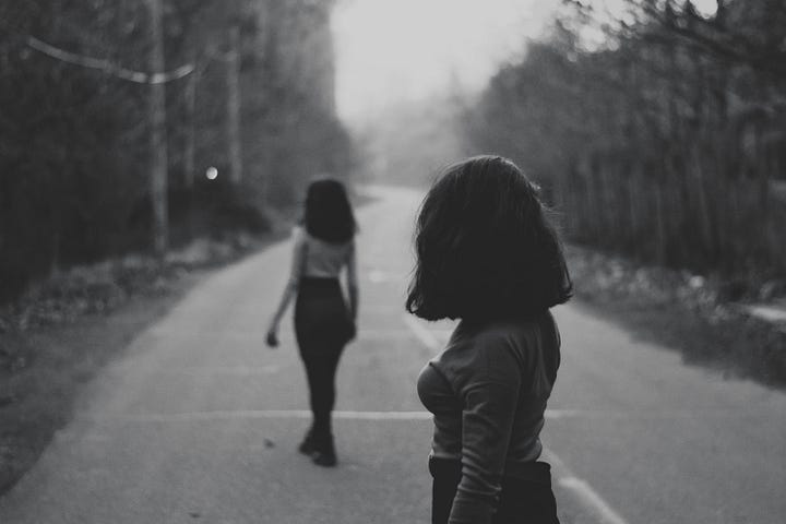 A young woman is looking back over her shoulder at another young woman who is walking away down an open road.