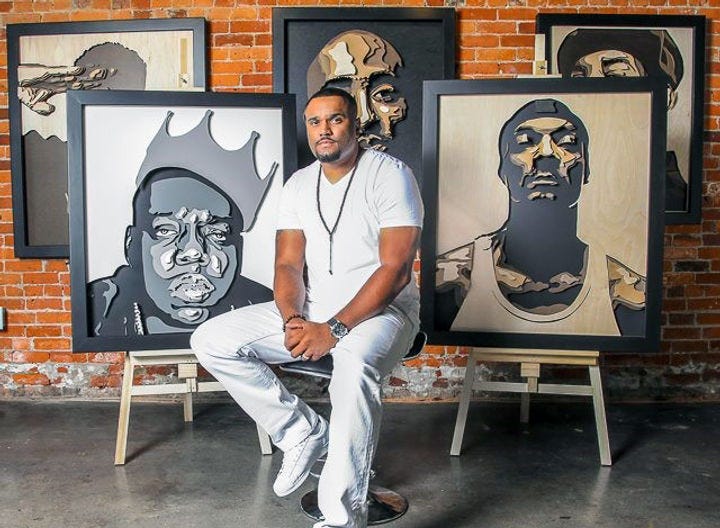 A Black man with short, dark natural hair and a short beard, wearing a white shirt and light jeans, sits in front of five paintings. All of the paintings are portraits of Black men.