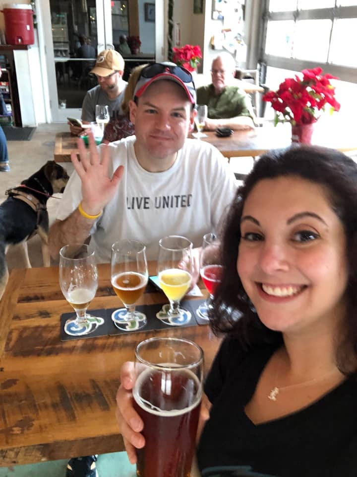 Andrew sits with a flight of beer in front of him, waving. I am taking the photo as a selfie holding a beer.