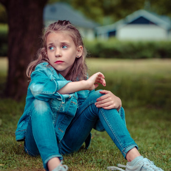 A sad-looking girl of about six or seven sits on the ground. She has big blue  eyes and light brown hair. She appears to be alone.