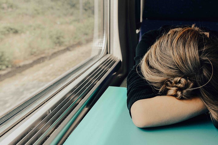 A person catching a nap while on a train or a bus, head on arms on the table in front of her.