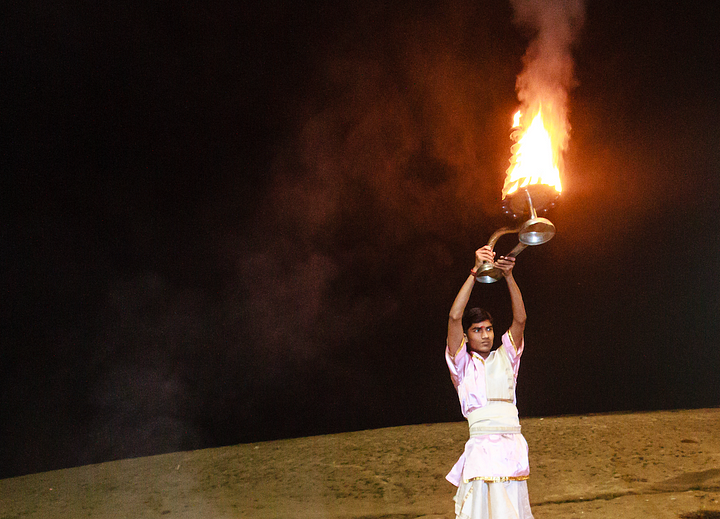 Against a background of the Ganges shore and a black night sky, a young man in ceremonial attire holds a tower of flame aloft.