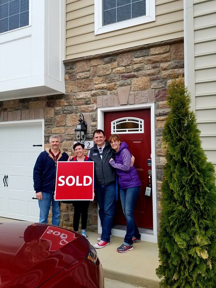 Photo of family, Dad, Daughter, Boyfriend, and Step-mom, outside a home with a brick exterior and red door. The daughter is holding a red sign that reads “SOLD” in white and upper case letters. The home is a town home, three levels with the upper levels having beige siding and a white pop-out on the second level. A red car is in the driveway in front of the family.