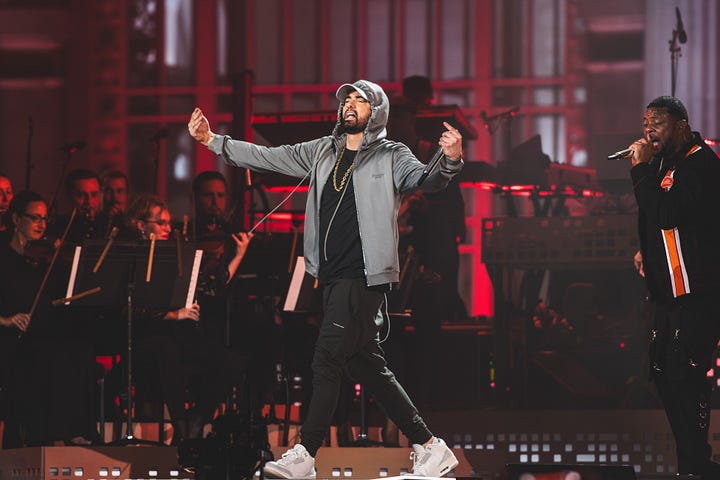 Eminem on stage, hands confidently spread wide to the audience, with a background singer and musicians behind him