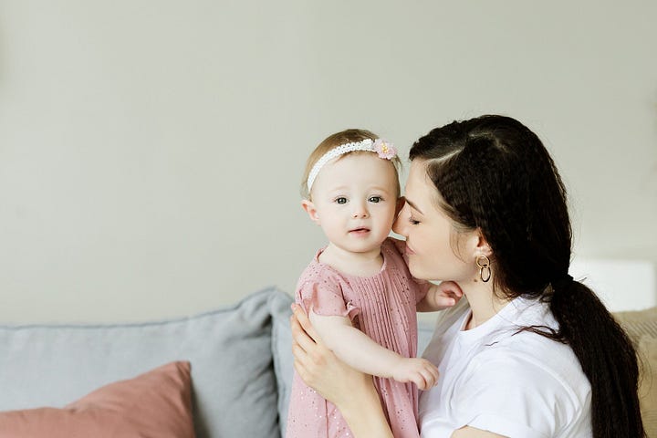 Mother with long dark hair in white shirt on couch closing her eyes and leaning into little girl looking straight at the camera.