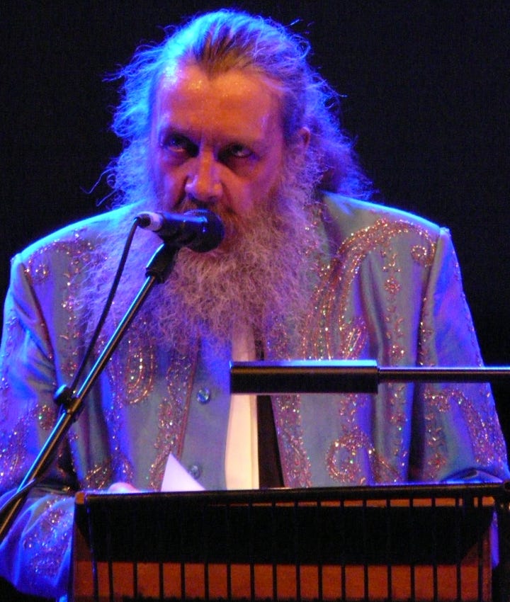 Author Alan Moore, an older man with brownish gray long hair and a scraggly beard, wearing a light colored blazer with swirling sparkling pattern. He’s standing at a lectern with a microphone, leaning in close and speaking.