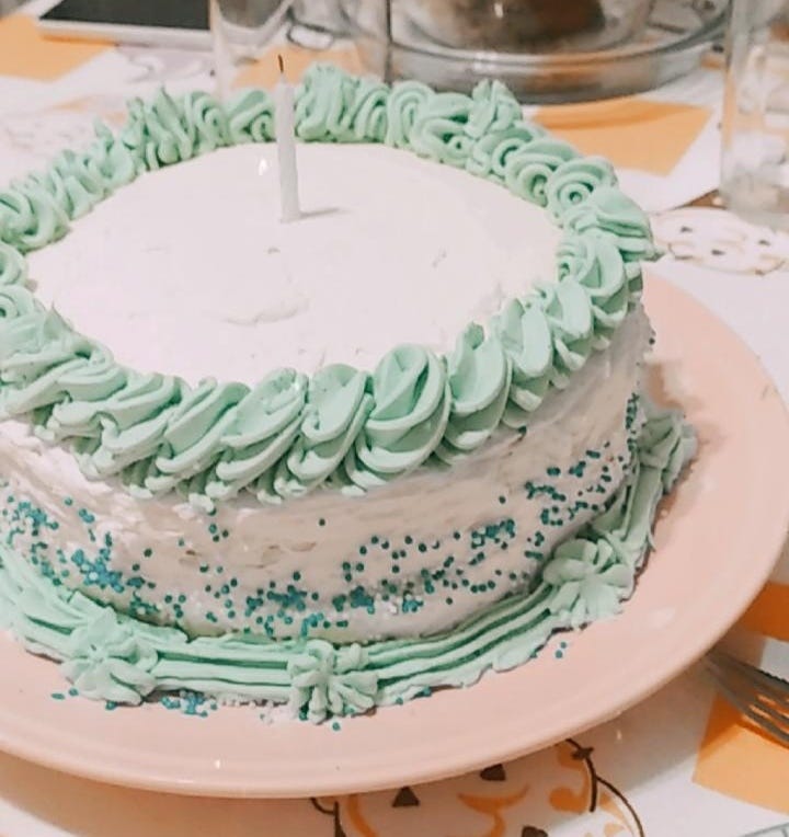 Foto de um bolo de aniversário com cobertura branca com detalhes azuis em um prato rosa claro e uma única velinha no meio