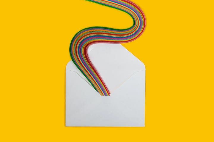 An open paper envelope on a yellow surface. A serpentine rainbow snakes its way out of the envelope.