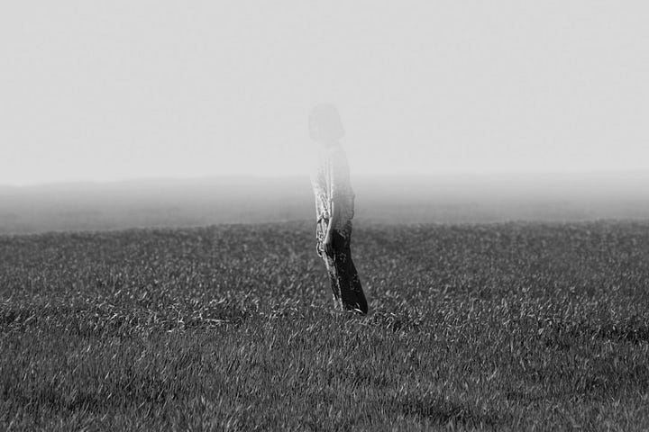 Black and white photo of person standing in field, a layer of fog is about 3 feet off the ground, making the upper portion of the person appear to fade and disappear.