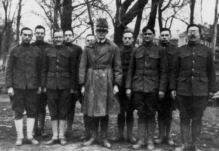 Black-and-white photo of a group of nine men in military uniforms standing outdoors in front of bare trees. The man in the center wears a long trench coat and hat, while the others wear standard-issue wool uniforms. Norbert Wiener, later known as the father of cybernetics, is standing on the far right.