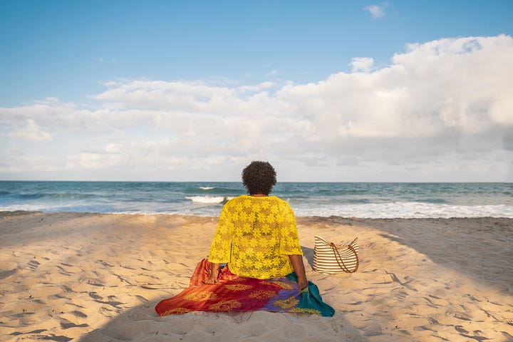Rear view of a fat Black woman sitting on a towel on the beach, watching the ocean.