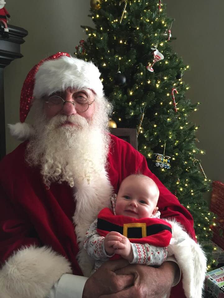 Bearded man with round gold frames sits in front of a Christmas tree in a Santa suit with a baby on his lap.