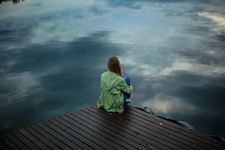 A young woman with her back to us, wearing a green rain jacket and jeans, sitting hugging her knees, on a wooden dock, looking out across still water reflecting a darkly clouded sky.