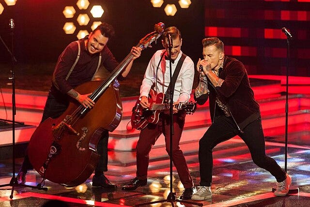 Three men singing and playing instruments on a stage with a dark red backdrop.