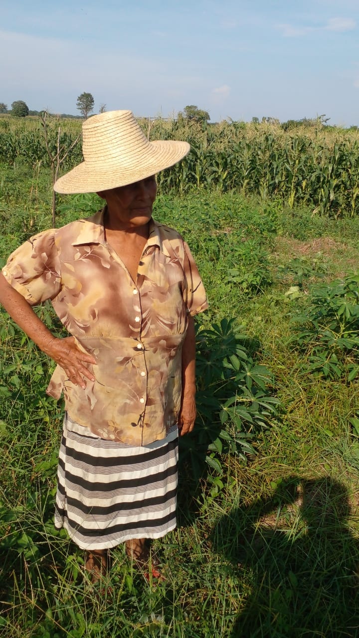 Una mujer de mediana edad está parada delante de su cultivo de maíz que se puede ver al fondo. La mujer lleva un sombrero que le cubre el rostro.