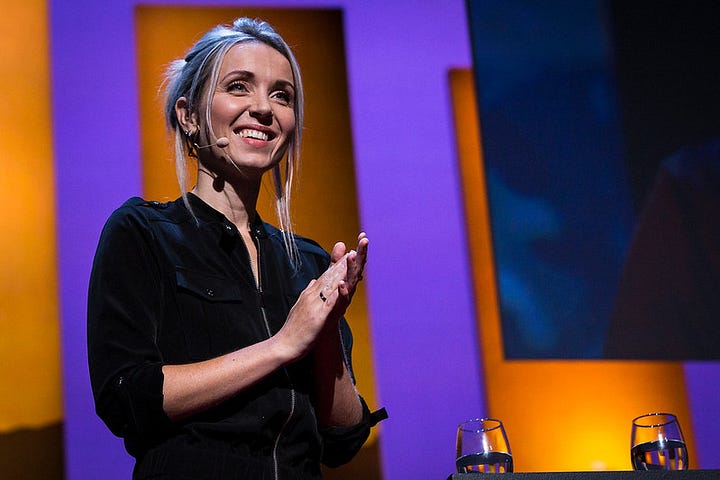 Thordis wears a black jumpsuit, hair up, smiling with hands clasped loosely in applause in front of backlite copper panels with purple in between, glasses of water and a black screen behind her.