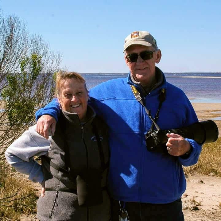 Carol and Russ smile together on a cool, sunny day at the beach. Russ holds a large camera with his arm around Carol.