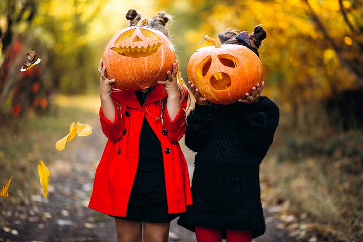 Two young girls dressed in autumn clothings holding pumpkins jack-o-lanters in front of their faces.