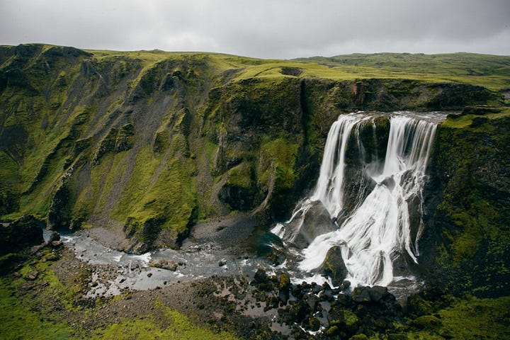A long green cliffside, with a section with overflowing water, creating a waterfall to a very rocky surface below.