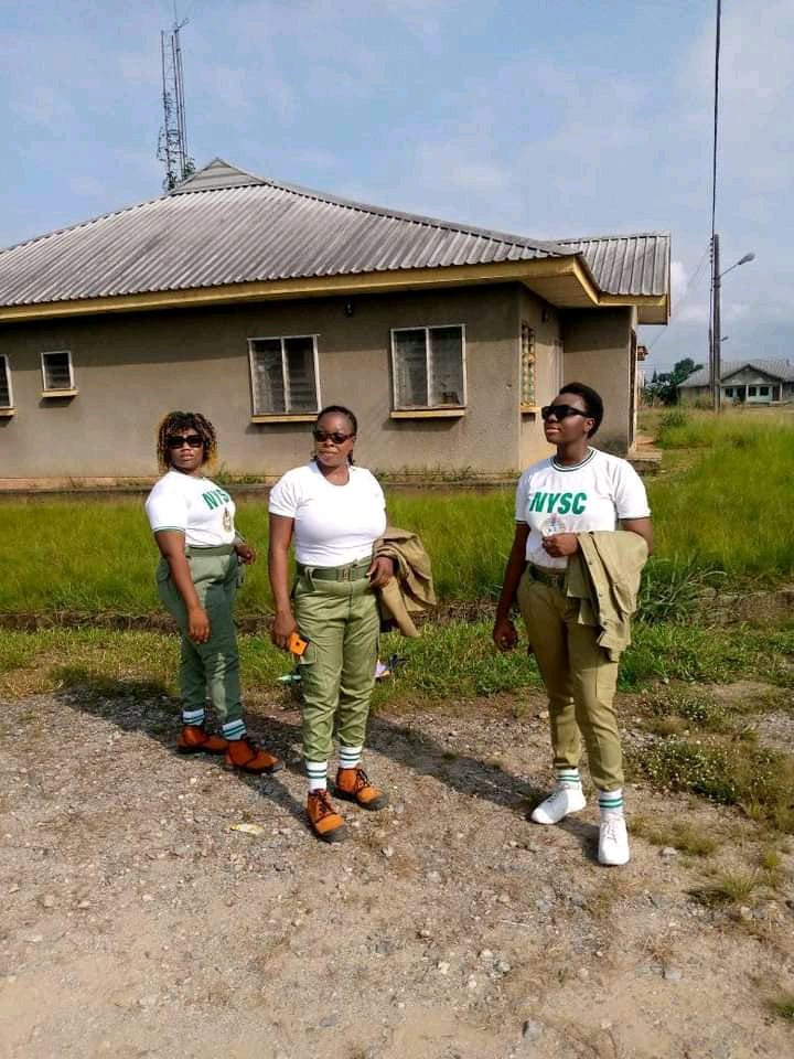 Three black big girls proudly wearing the Nigerian National Youth Service Corps uniform, standing in front of an old apartment with dark shades, posing for the camera.