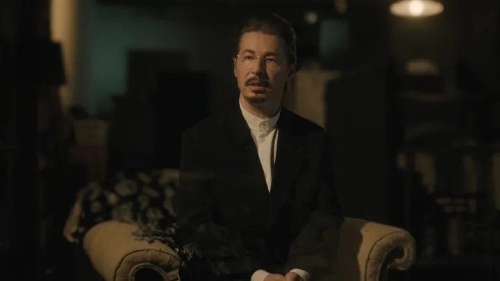 Actor Edoardo Ballerini is shown in a black suit, white shirt, thin wire-framed glasses, with a moustache and a goatee, appearing slightly transparent, sitting in an armchair as his character, Ray.