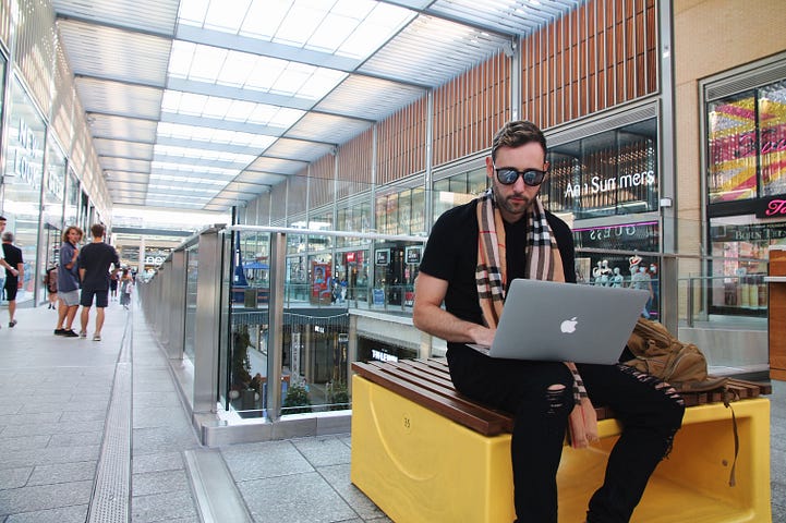A man sat on a seat in a shopping centre. He has an Apple laptop open on his lap. He is wearing sunglasses and a has a scarf around his neck. Is he replying to negative comments on his Medium stories?