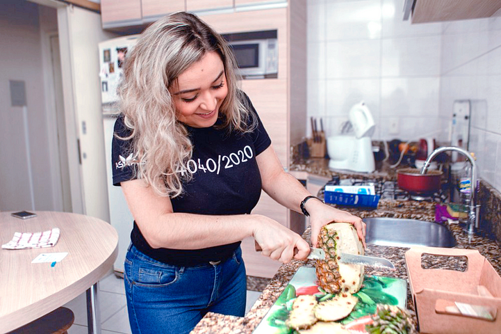 Bianca Delabenetta, líder da equipe de Empreendedores no Asaas, sorri enquanto descasca um abacaxi em sua cozinha. Ela possui cabelos loiros e veste uma camiseta preta.