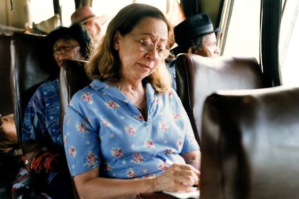 Mulher branca, cabelos castanhos claros, está sentada no ônibus de viagem e está escrevendo em um papel. Ela veste uma camisa azul com estampa de flores na cor rosa.