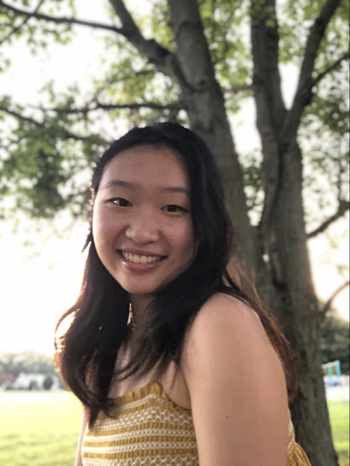 A smiling person stands outside in front of a tree.