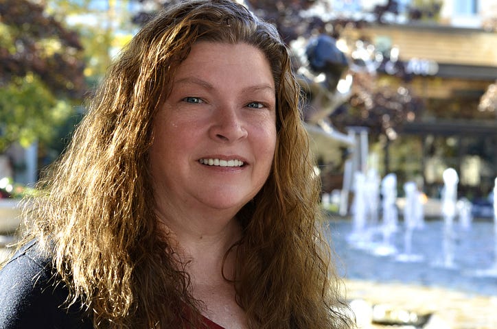 Photo of author JoEllen Claypool with shoulder-length brown hair with a burgundy shirt and black cover shirt with fountains in the background.