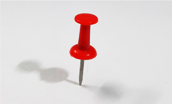 a single red thumbtack on a pinned to a slightly grey background. Object: Red Thumbtack, Action: Pinned on a flat surface, Context: Very light grey paper kept on horizontal surface