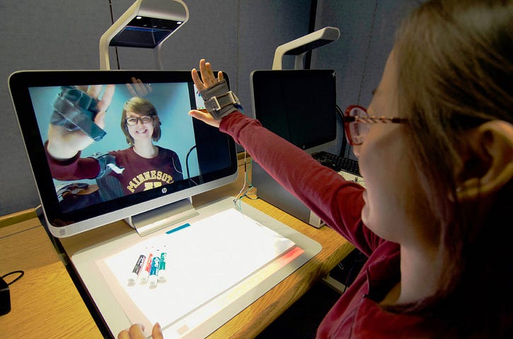 A woman raises her hand towards a webcam during a videochat with a friend. Her hand is encased in a cloth device with shape memory alloy springs.