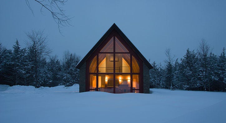 A well lit, wooden house sits on a snowy field in the evening.