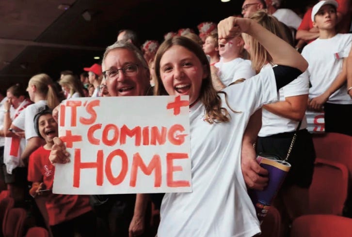 photo of happy girl pointing to handmade sign saying “It’s Coming Home” held up by her father