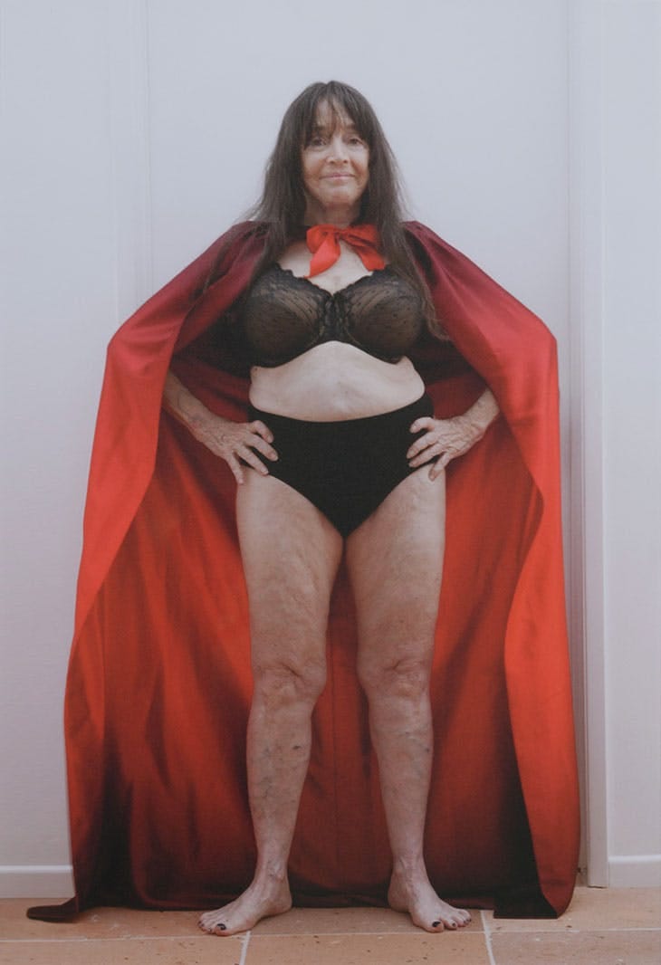 Self Portrait of Eleanor Antin, she’s in her 80s, wearing a black bra and underwear and wearing a red cape, hands on hips.