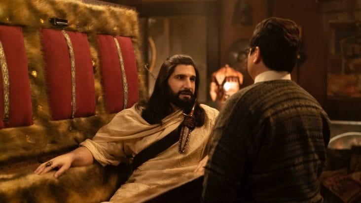 Nandor The Relentless (Kayvan Novak) in a fur-lined coffin with the lid propped open, slightly sitting up and talking to his Familiar, Guillermo (Harvey Guillen).