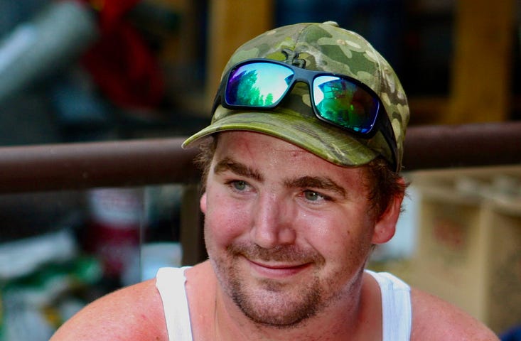 The face of a young man with a smile on his face, a small beard. He’s wearing a camouflage cap with sunglasses on top