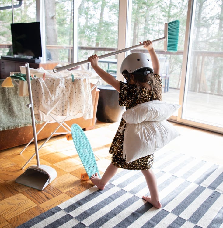 A child wearing a skateboard helmet with a cushion strapped to her back holds up a household brush. The room she’s in has wooden floors and wide windows, and also contains a sofa, TV, washing stand with washing, and a blue skateboard.