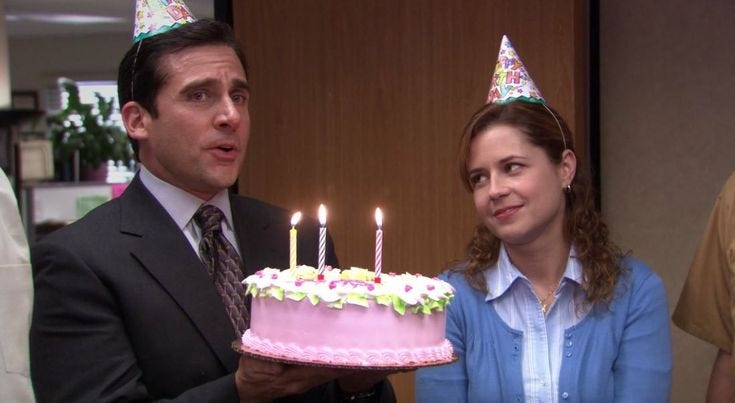 No episódio “Dinheiro” da temporada 4 de “The Office”, Michael Scott celebra com um bolo de aniversário rosa, segurando-o com entusiasmo ao lado de Pam Beesly. Ambos vestem chapéus de festa e exibem expressões de alegria, representando o clima de camaradagem no escritório da Dunder Mifflin. Pam, sorrindo, olha para o bolo decorado com flores de glacê e três velas acesas, enquanto Michael está cantando anunciando a celebração em um ambiente de escritório.