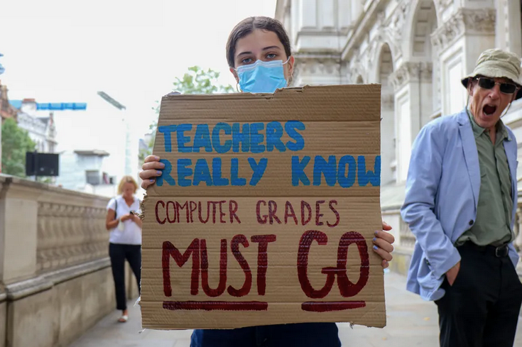 Woman wearing a mask with a cardboard sign: ‘Teachers really know computers grades MUST GO’