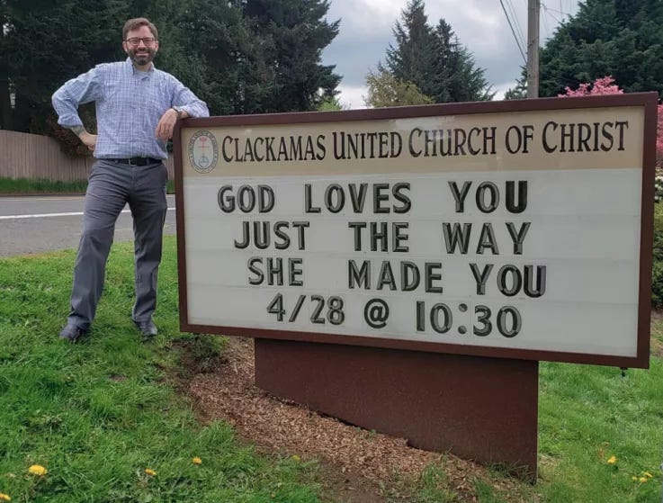 A white bearded person with glasses stands proudly next to a church sign reading “God loves you just the way she made you”