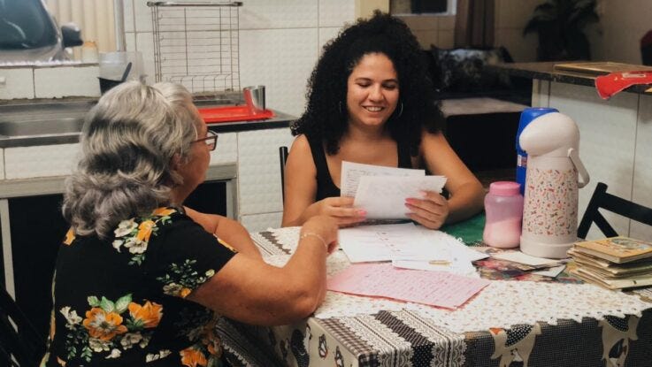 No canto esquerdo da imagem vemos a avó de Maysa sentada à mesa com sua neta, que sorri enquanto lê um dos textos escritos à mão pela avó. Elas estão sentadas na mesa da cozinha da avó de Maysa, em Coruripe.
