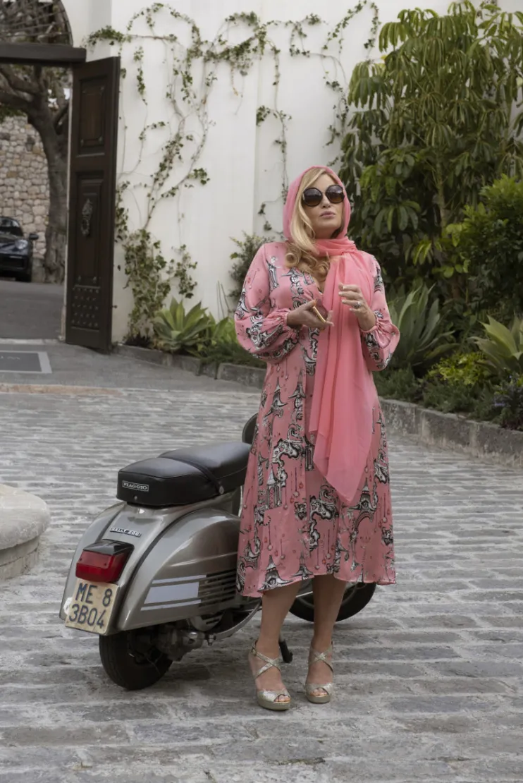 A still from The White Lotus, with Jennifer Coolidge wearing a pink headscarf and dress and large sunglasses, standing in a courtyard in front of a Vespa.