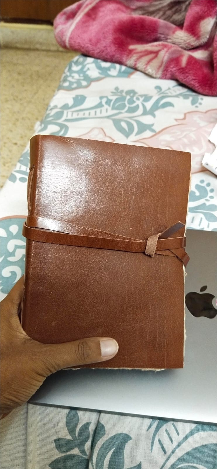 A hand holding a notebook with a leather cover. Beside it, a laptop sits on the bed.